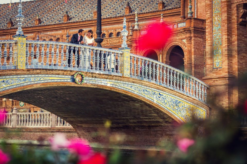 A couple dressed for a wedding stands on an ornate bridge with blue and white tiles.