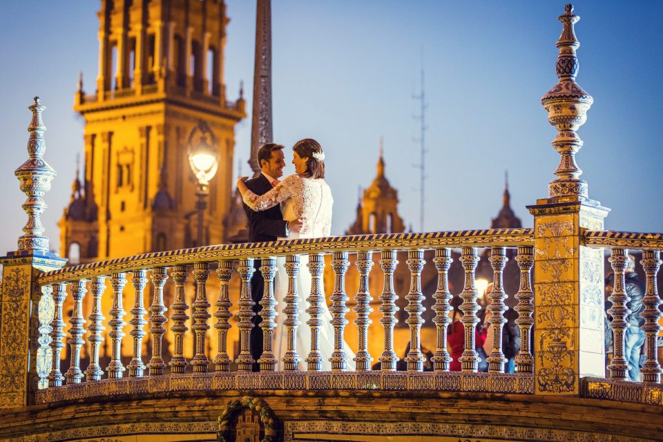 A couple dressed for a wedding dance in an embrace on an ornate bridge with tiled decoration and a decorative balustrade.
