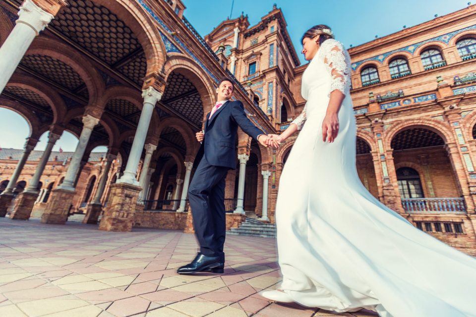 A couple dressed for a wedding walk hand in hand in front of a historic brick building with arches.