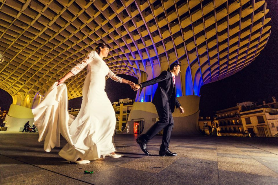 A couple dressed for a wedding walk hand in hand through a square at dusk.