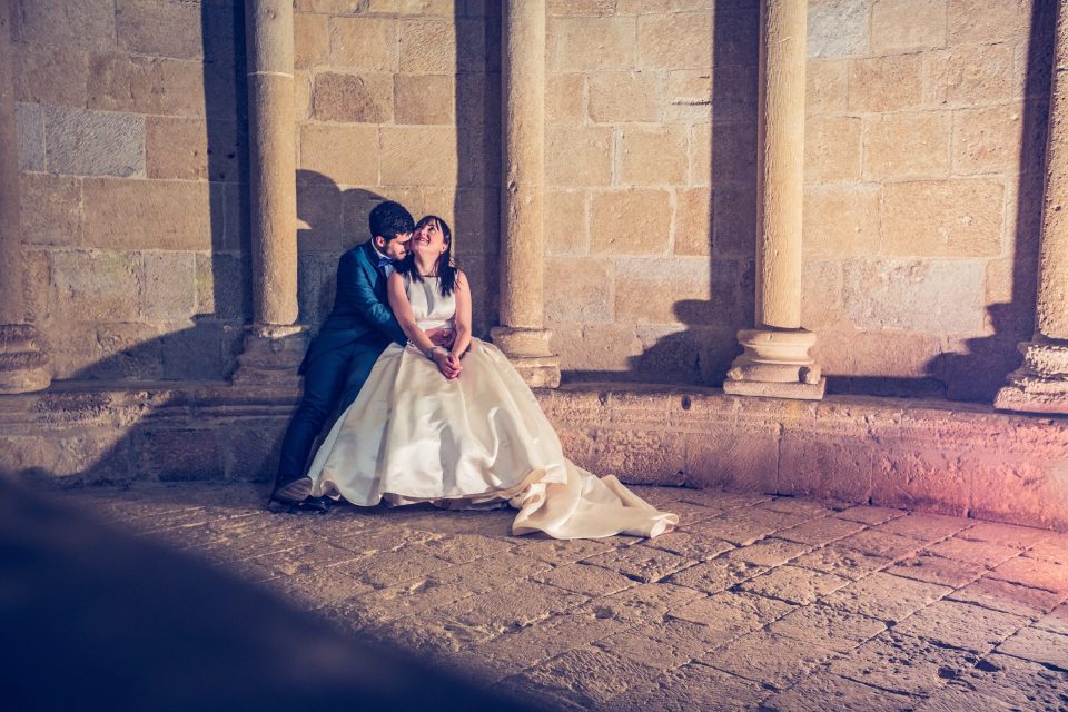 A couple dressed for a wedding is seated beside a stone wall with columns.