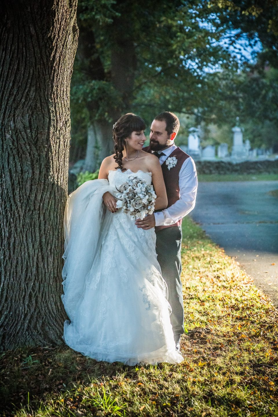 A couple dressed for a wedding pose outdoors beside a large tree.