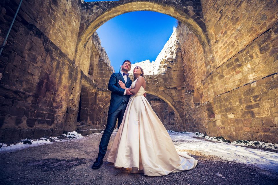 A couple dressed for a wedding pose beneath a large stone arch.