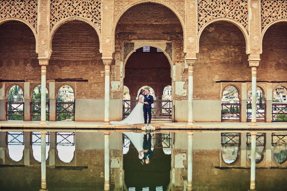 Una pareja vestida para una boda posa bajo una galería histórica con arcos, columnas y celosías ornamentadas.