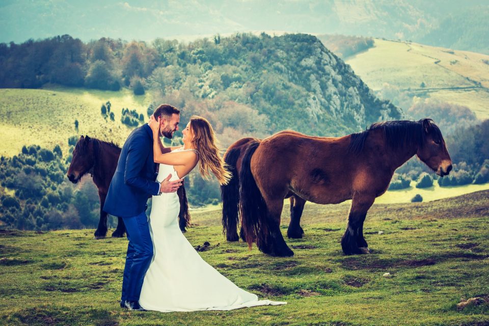 Una pareja vestida para una boda posa en un prado de montaña: él lleva un traje azul y ella un vestido blanco largo.