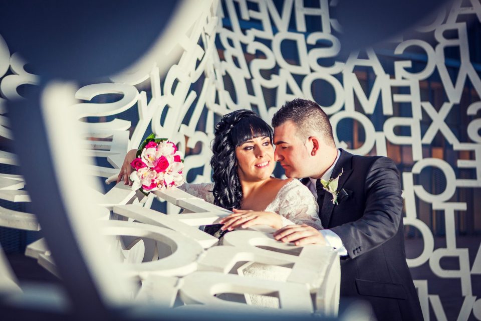 Una pareja vestida para una boda posa junto a una estructura decorativa blanca formada por letras y formas geométricas.