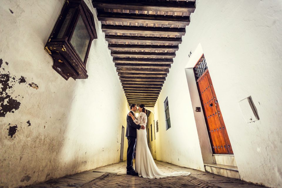 A couple dressed for a wedding embrace and kiss in a narrow passageway with white walls and a cobbled floor.