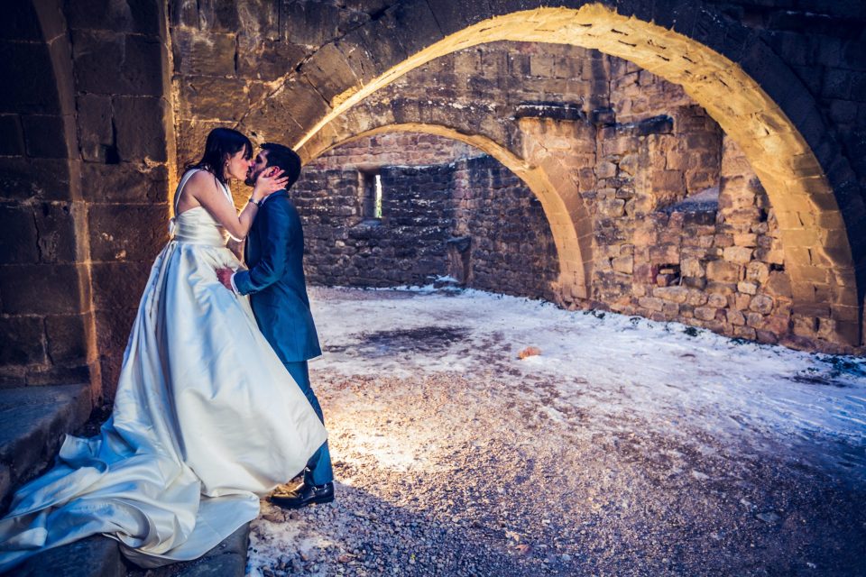 A couple dressed for a wedding kiss beneath a large stone arch in a historic brick-and-masonry space.