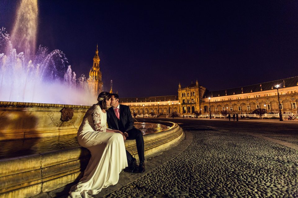 A couple dressed for a wedding kiss while seated beside a fountain lit up at night.