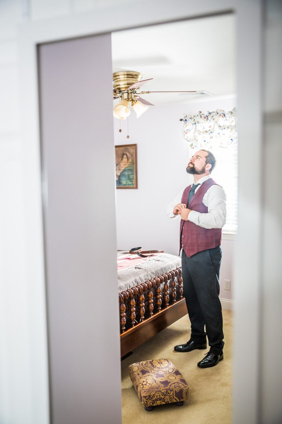 A bearded person gets ready in a room, seen through a doorway.