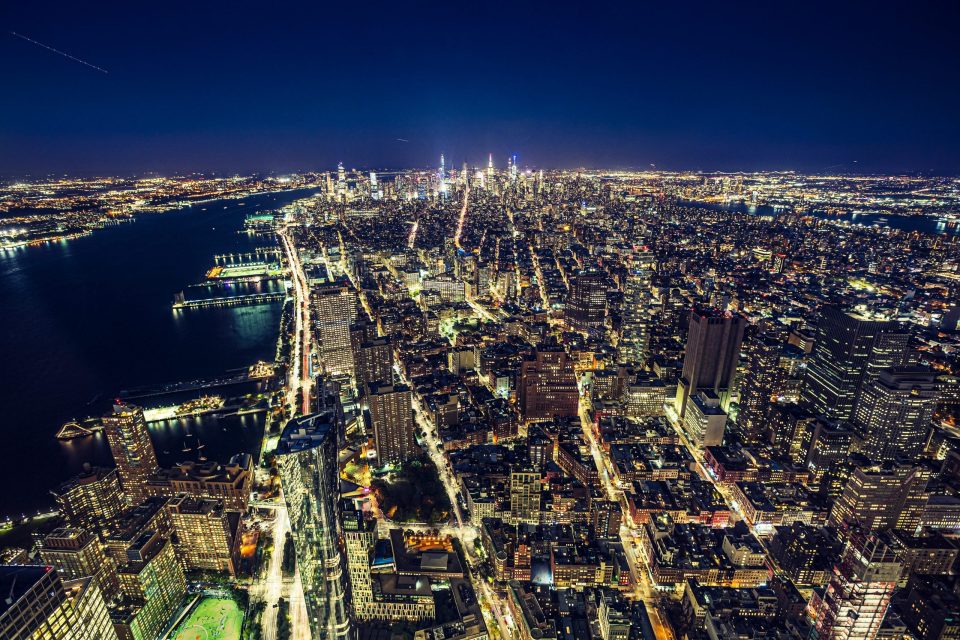 This is a night-time aerial view of a large coastal city, with numerous illuminated buildings and grid-patterned avenues.