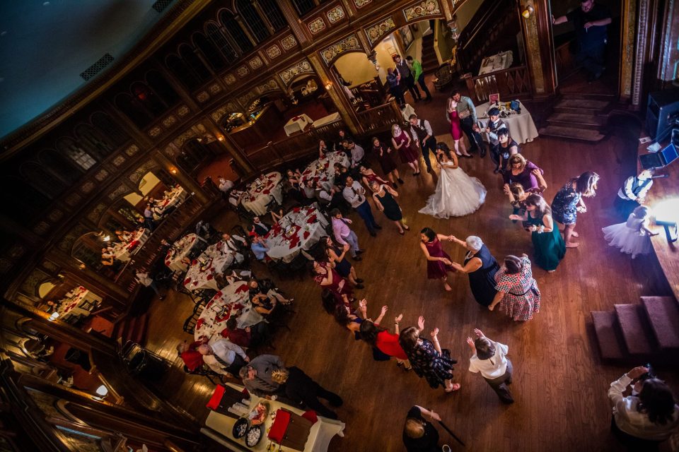 An elevated, slightly tilted view shows a wedding celebration in an ornately decorated wooden hall.