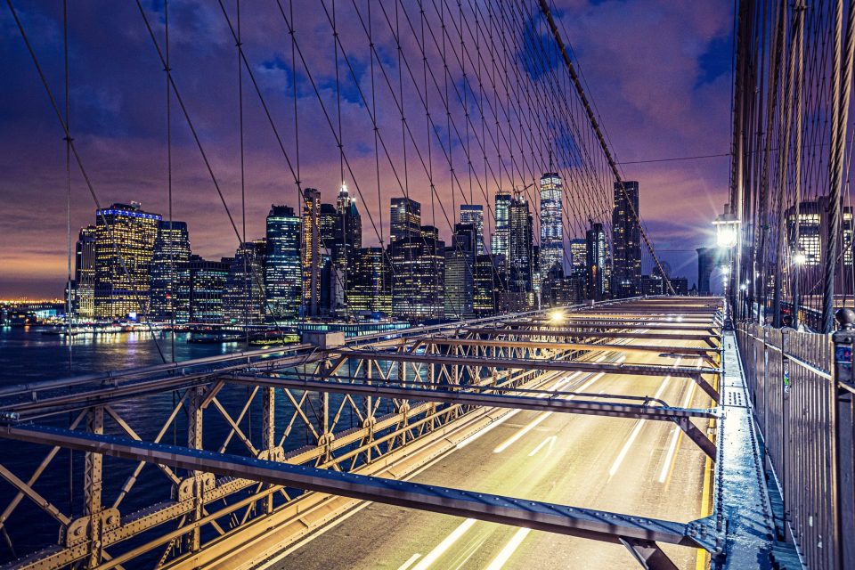 Night view of an illuminated suspension bridge, with cables and metal structure in the foreground.