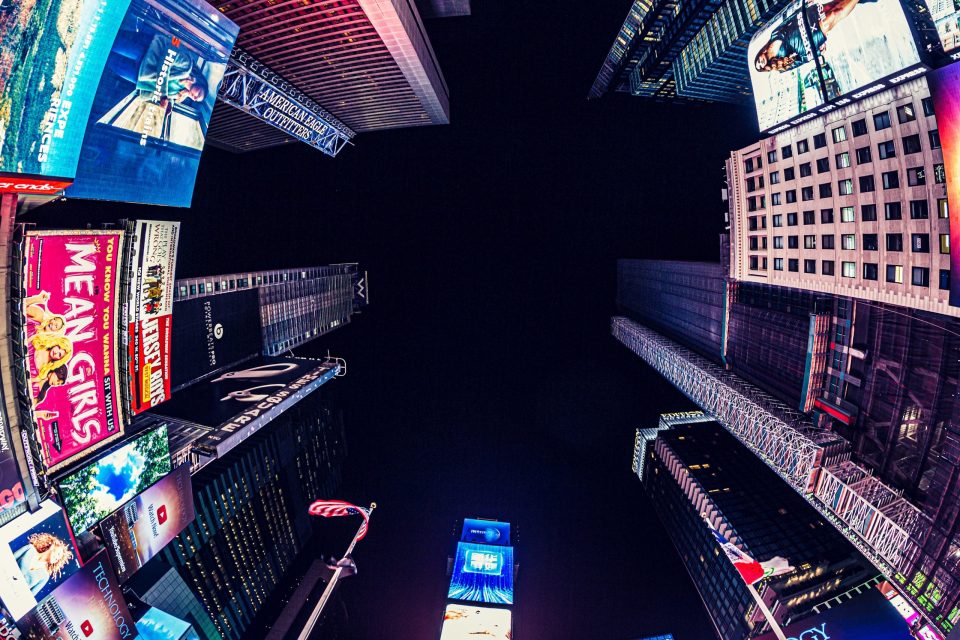 A low-angle night view of an urban square surrounded by tall buildings and large illuminated advertising screens.