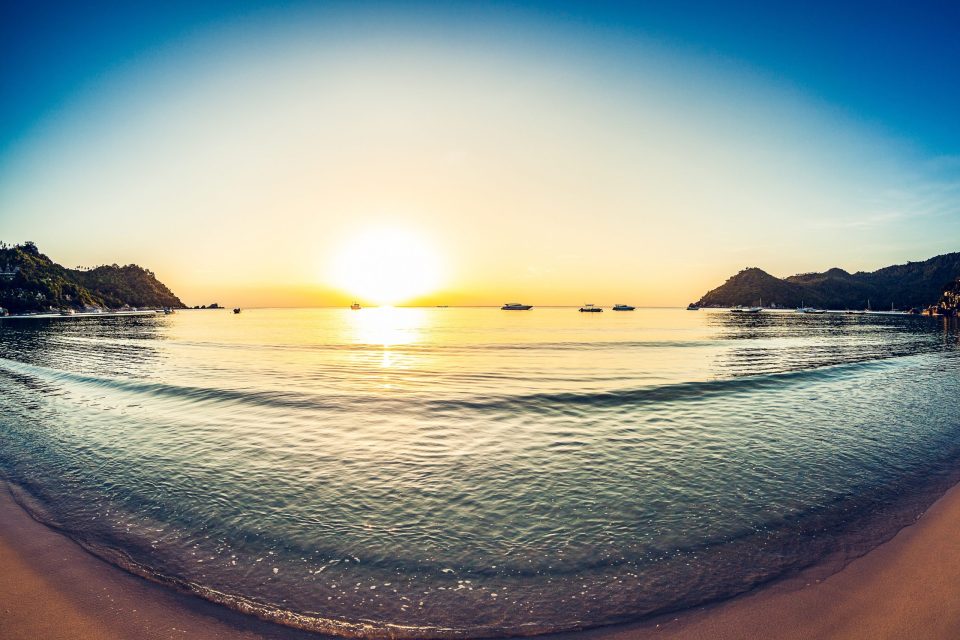 A panoramic view of a bay at sunset, with the sun low on the horizon and calm water with golden reflections.