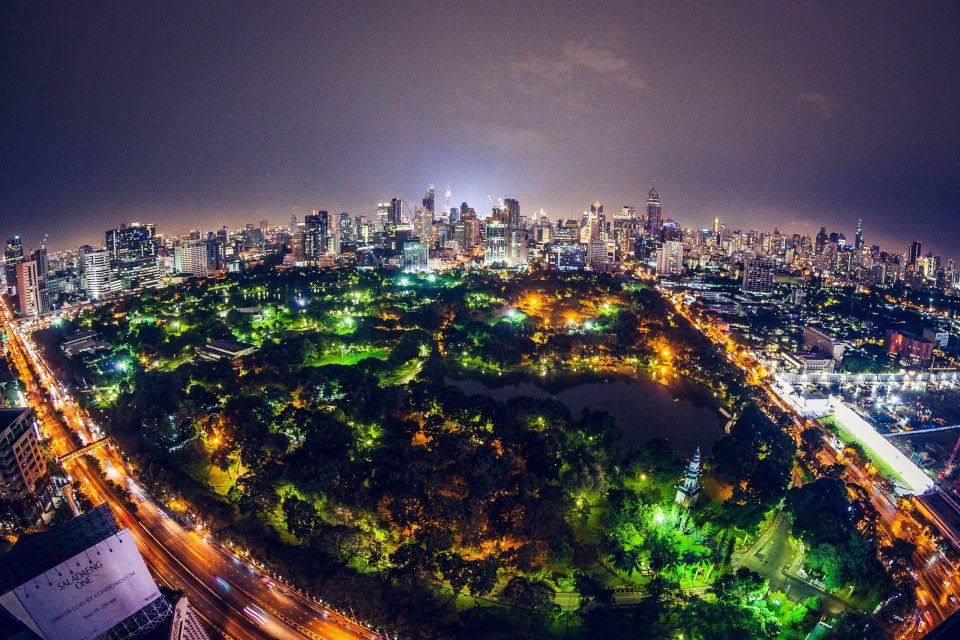 A night-time panoramic view shows an illuminated city, with a large wooded park and a pond in the foreground.