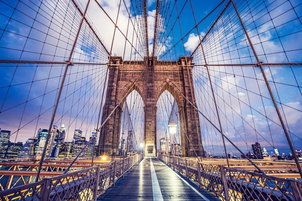 A symmetrical view of Brooklyn Bridge from the pedestrian walkway, with its cables and stone arches in the foreground.
